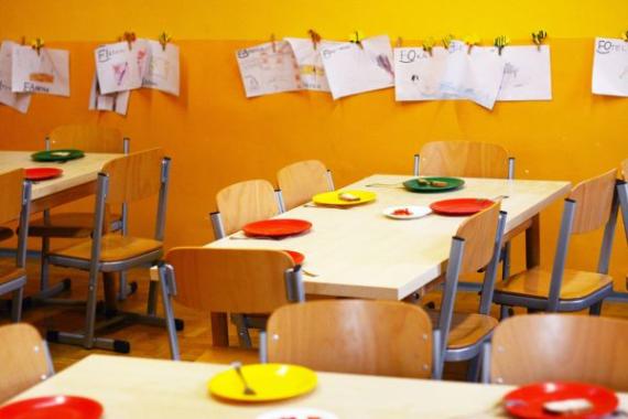 Nell'immagine: Stanza di scuola con muri arancioni. Ci sono tavoli con sedie di legno e piatti colorati. Sul muro sono attaccati con delle mollette i disegni dei bambini. 